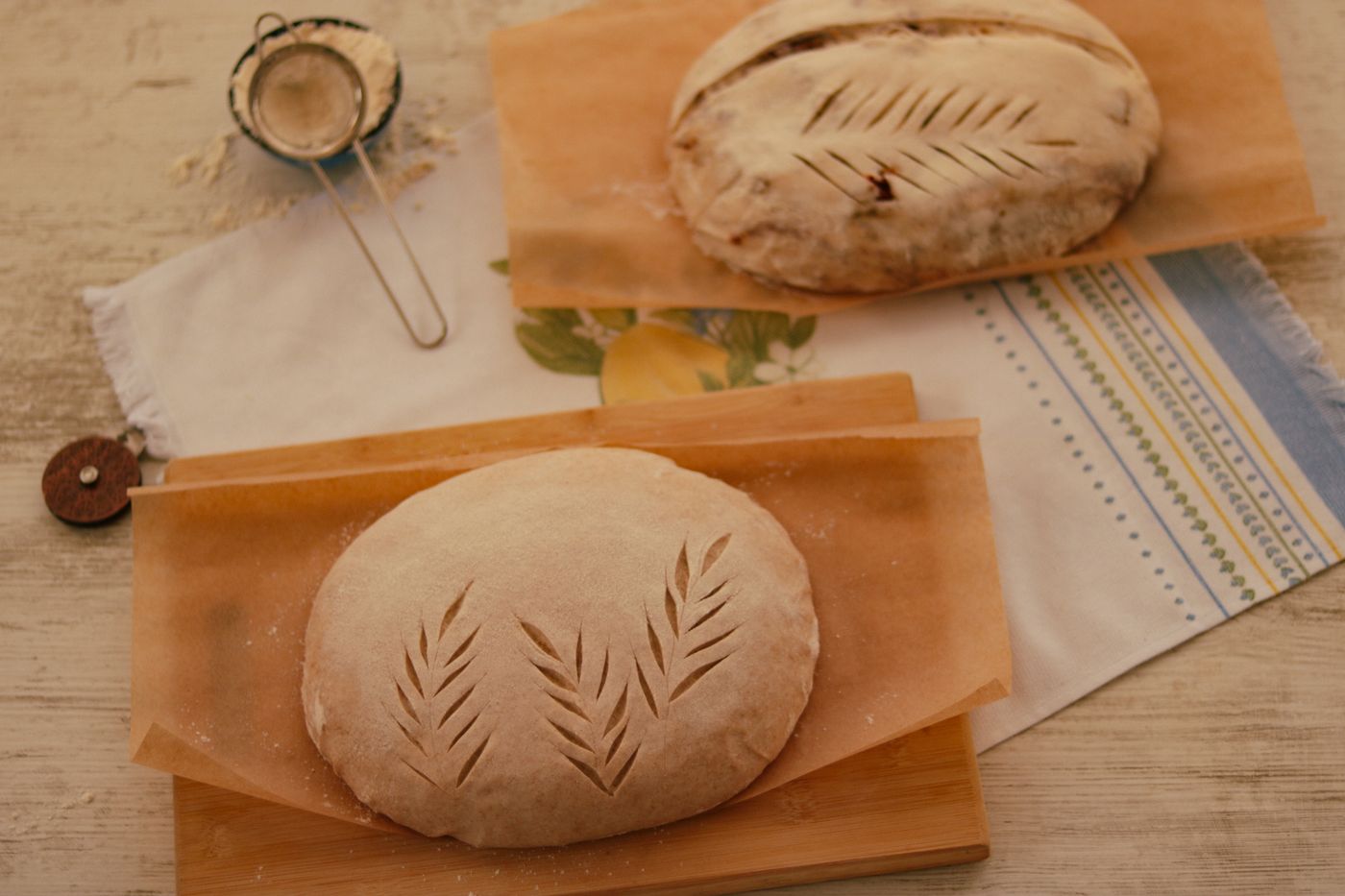 Two shaped dough loaves on parchment paper, one with a leaf pattern scored into the top