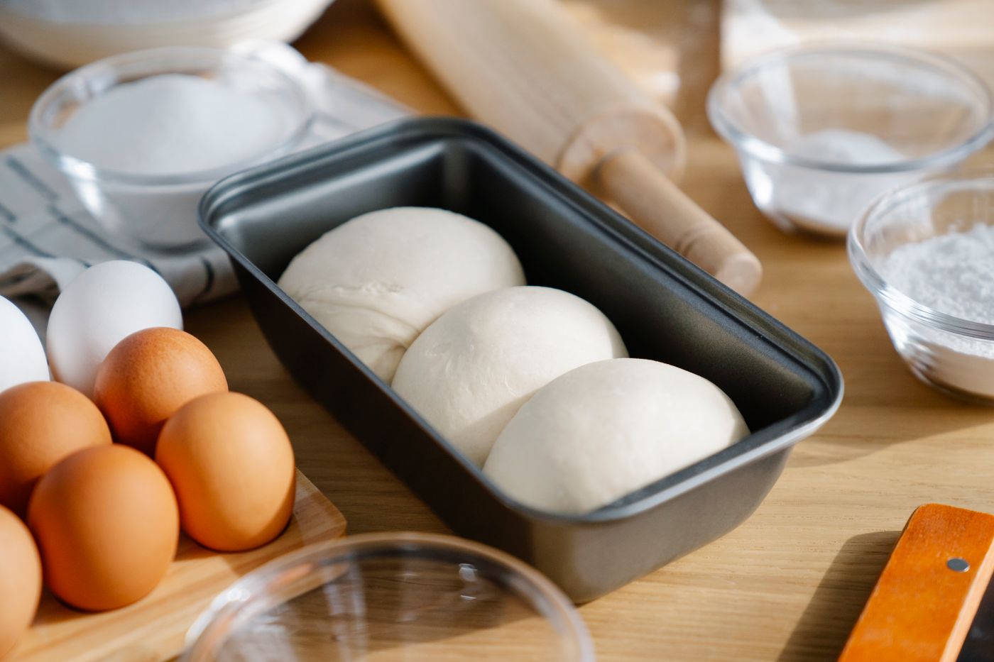 Flour, eggs, water and a loaf pan with dough balls laid out on a wooden kitchen table